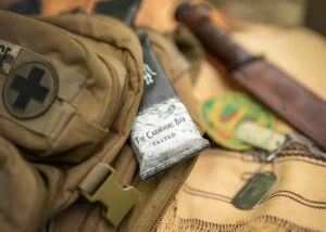 A close-up of a carnivore bar with the flavor ‘Salted’ placed on top of a tactical backpack next to a knife and dog tags, suggesting readiness for outdoor activities or survival scenarios.